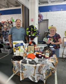 Photo of Mr. & Mrs. Beth Roberts the winner of our September Raffle drawing. The picture shows a round table with a table cloth displaying miscellanious merchandise that was raffled off in cluding cast iron cookware, towels, housewares, jams, toys, art, rare coin, and gift cards with a total value over $600.00
